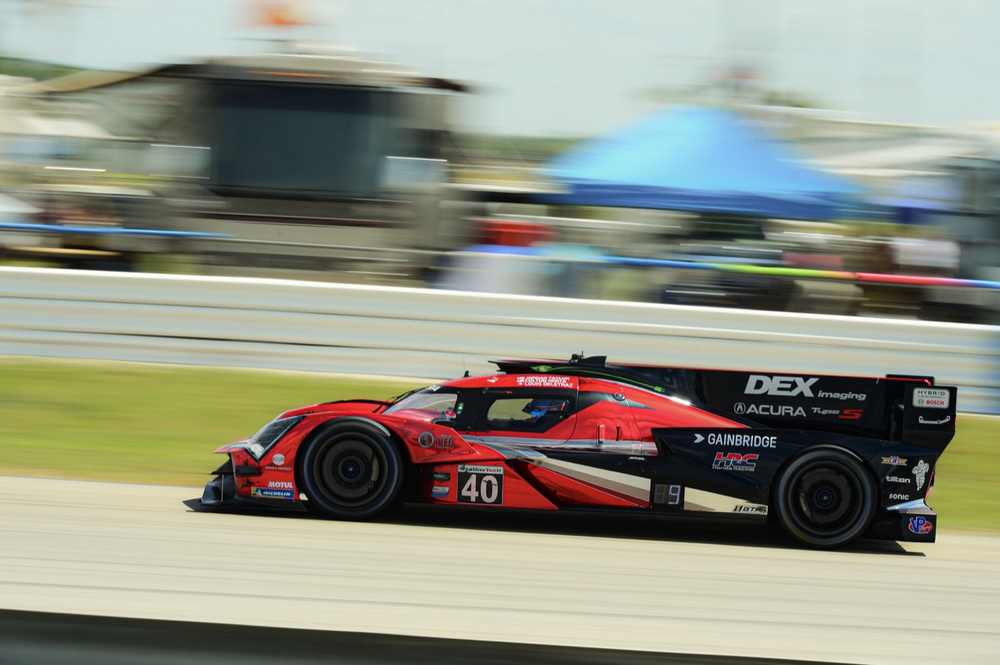 Red and black Acura ARX-06 prototype number 40 at speed in daylight, with blurred hospitality tents behind