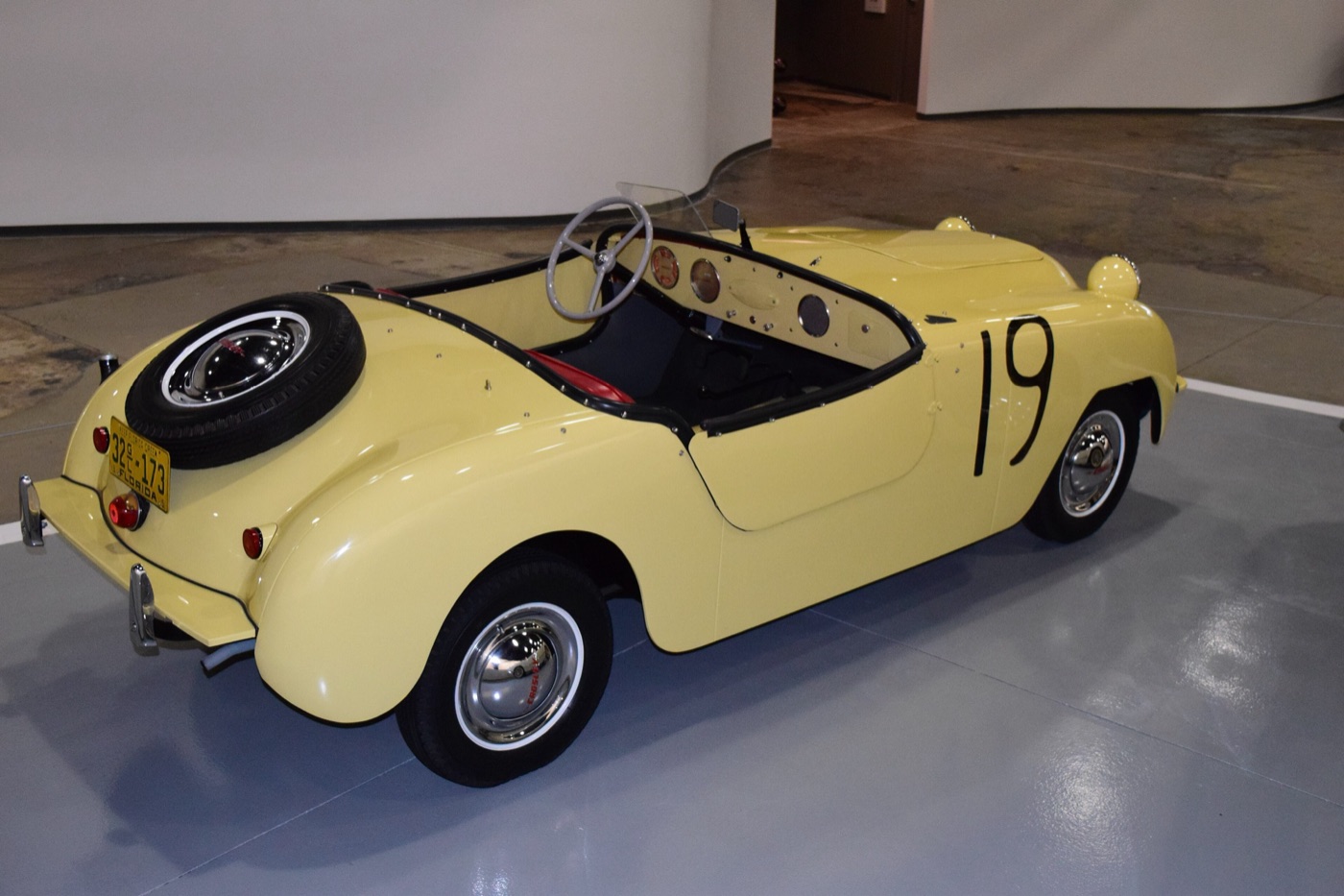 Pale yellow Crosley Hot Shot roadster with race number 19 and a spare wheel on its tail, on a museum floor