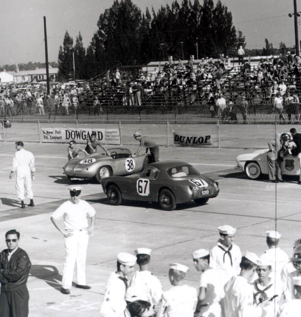 Black and white photograph of mechanics pushing small sports cars numbered 38 and 67 in front of a packed wooden grandstand, with sailors in white uniforms in the foreground