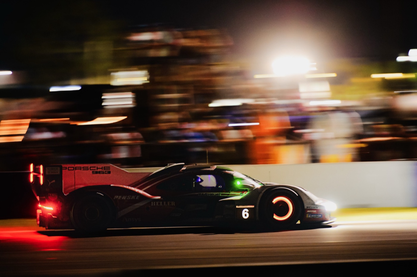 Porsche 963 prototype number 6 at speed under floodlights, its brake discs glowing, with a blurred crowd behind