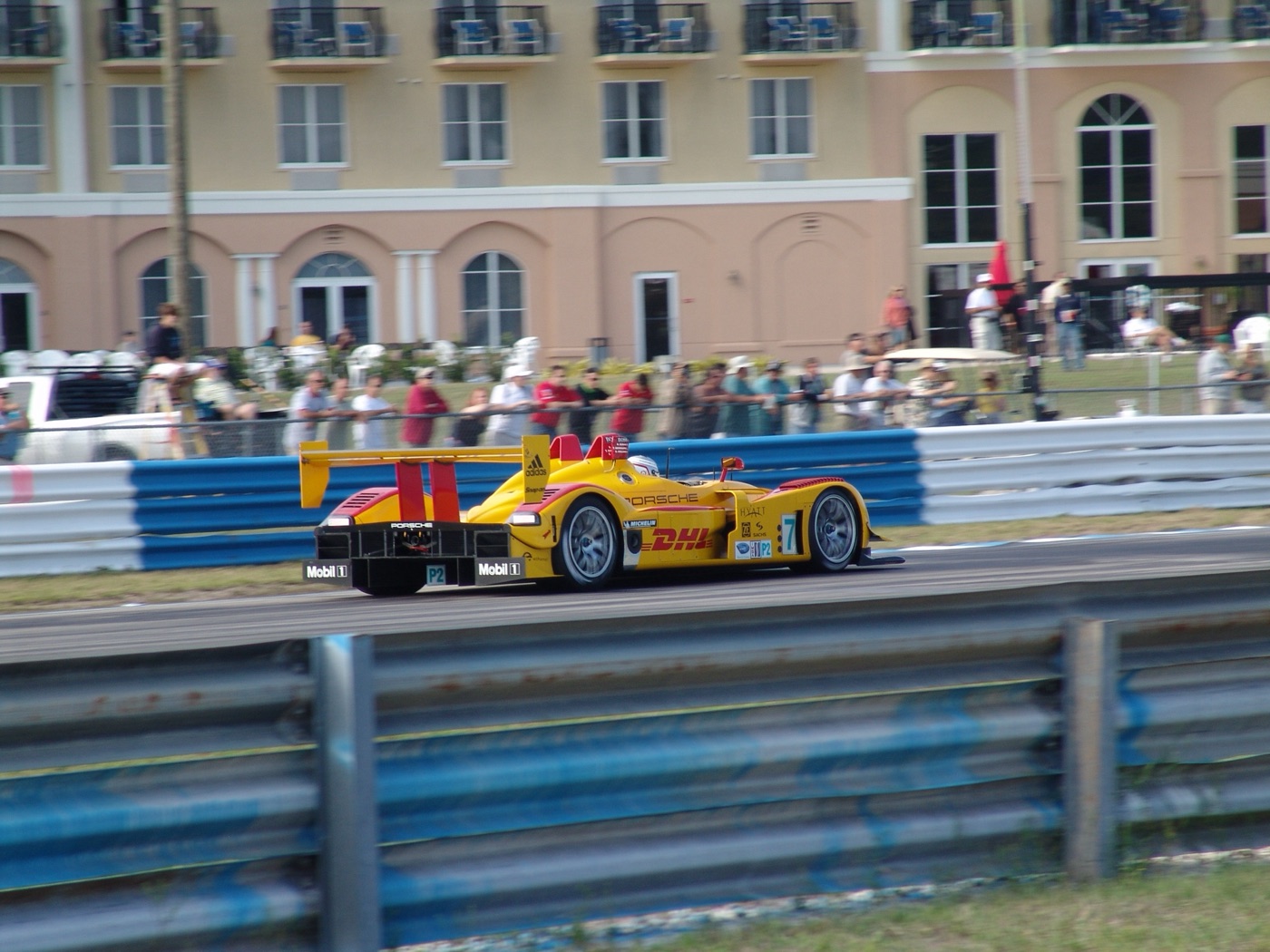 Yellow and red Porsche RS Spyder prototype number 7 on track in front of a pink hotel building with spectators along the fence