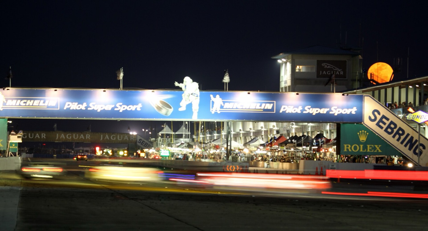 Light trails of race cars passing the pits under the Michelin bridge at night, with the Sebring sign and a full moon rising behind the tower
