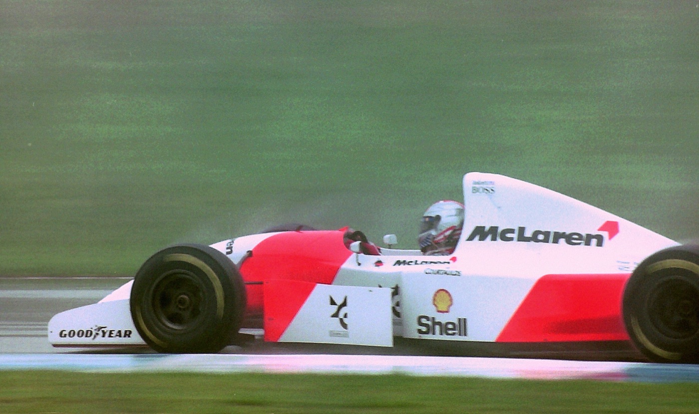 White and red McLaren Formula One car with a silver helmeted driver on a wet track, spray behind