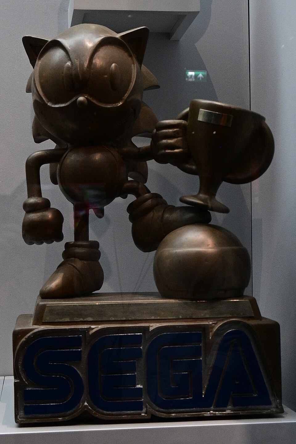 Bronze statue of Sonic the Hedgehog holding a cup, one foot on a ball, on a base with the word SEGA in blue, inside a glass case