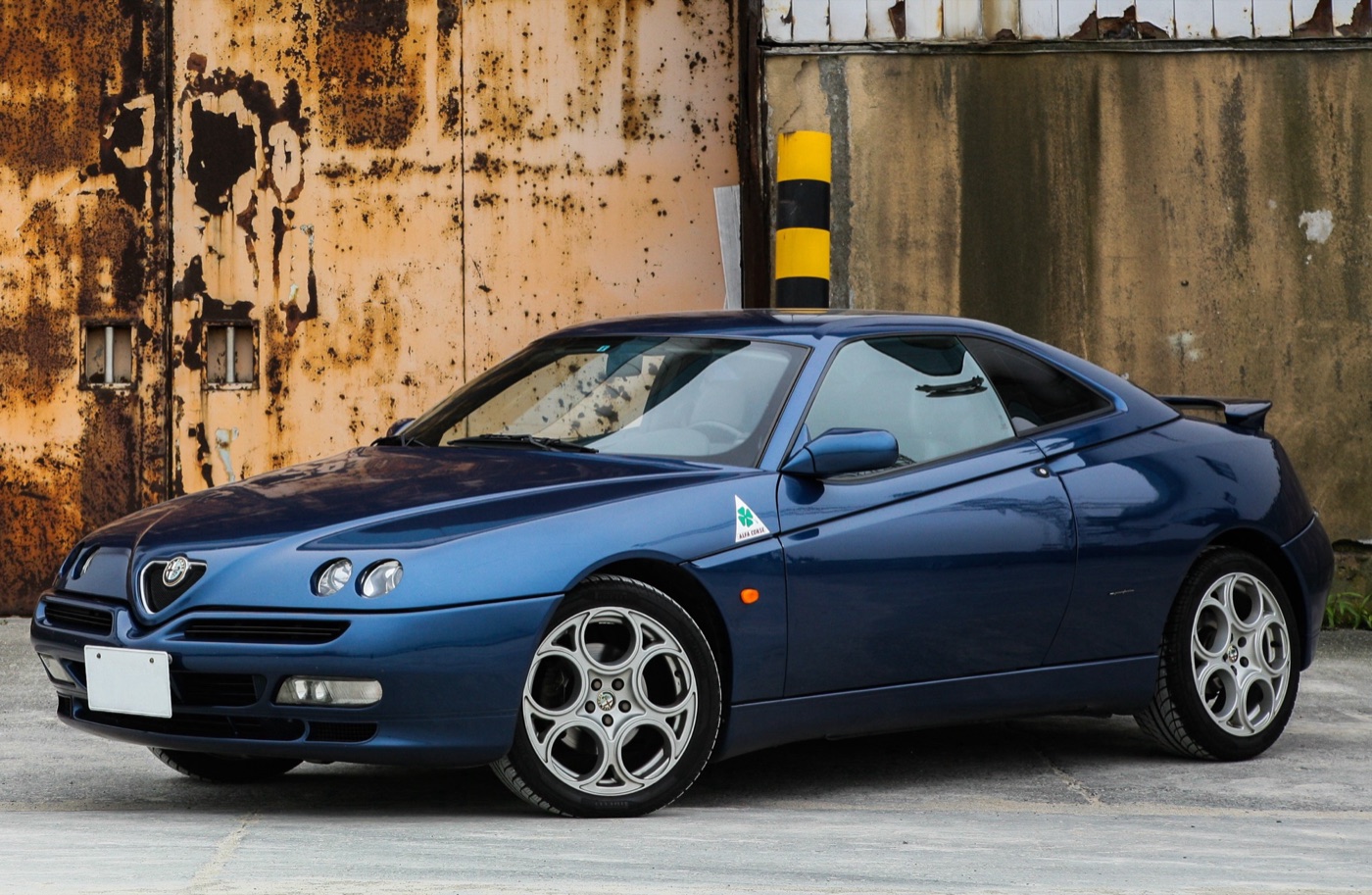 Blue Alfa Romeo GTV coupé with five-hole alloy wheels and twin round headlamps, parked in front of a rusted steel wall