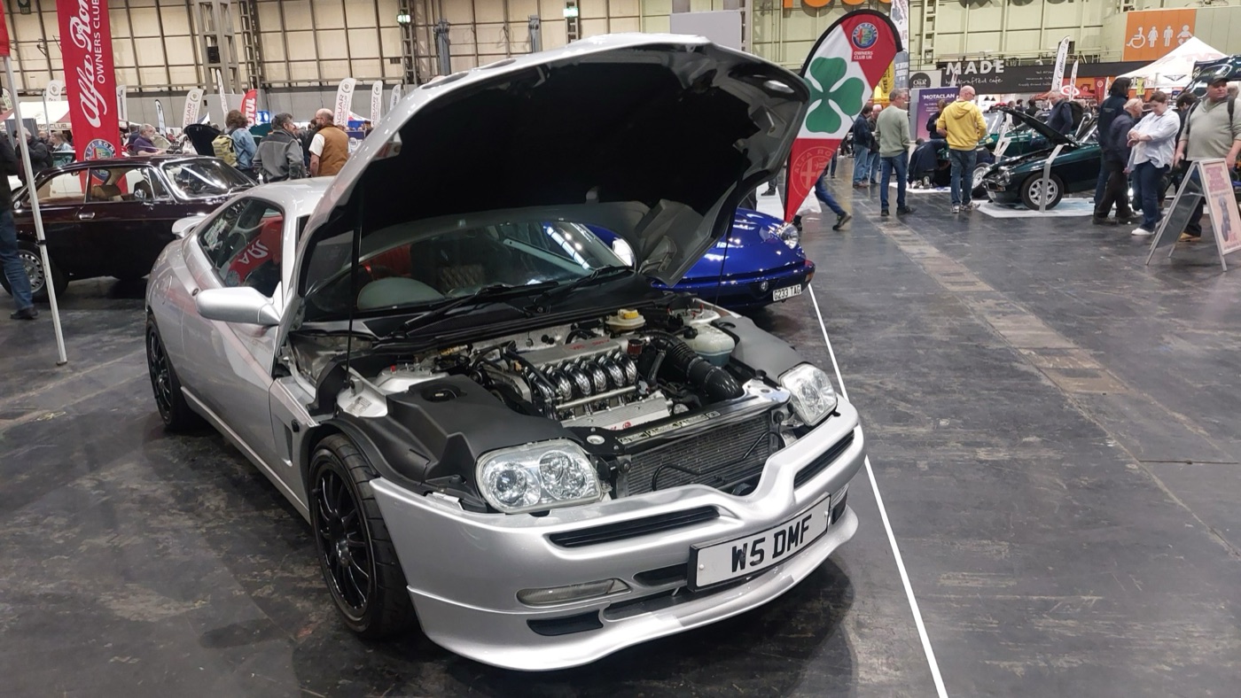 Silver Alfa Romeo GTV with its one-piece bonnet raised, showing the V6 engine and the fixed headlamps left in place on the body