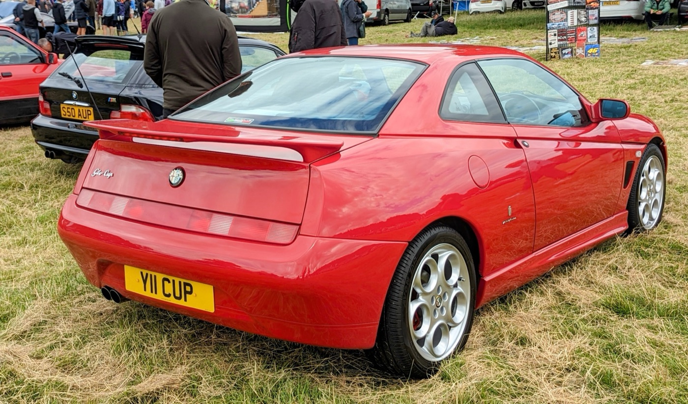 Red Alfa Romeo GTV Cup seen from the rear three-quarter on grass at a car show, with a large rear wing, side skirts, a GTV Cup badge and a UK number plate