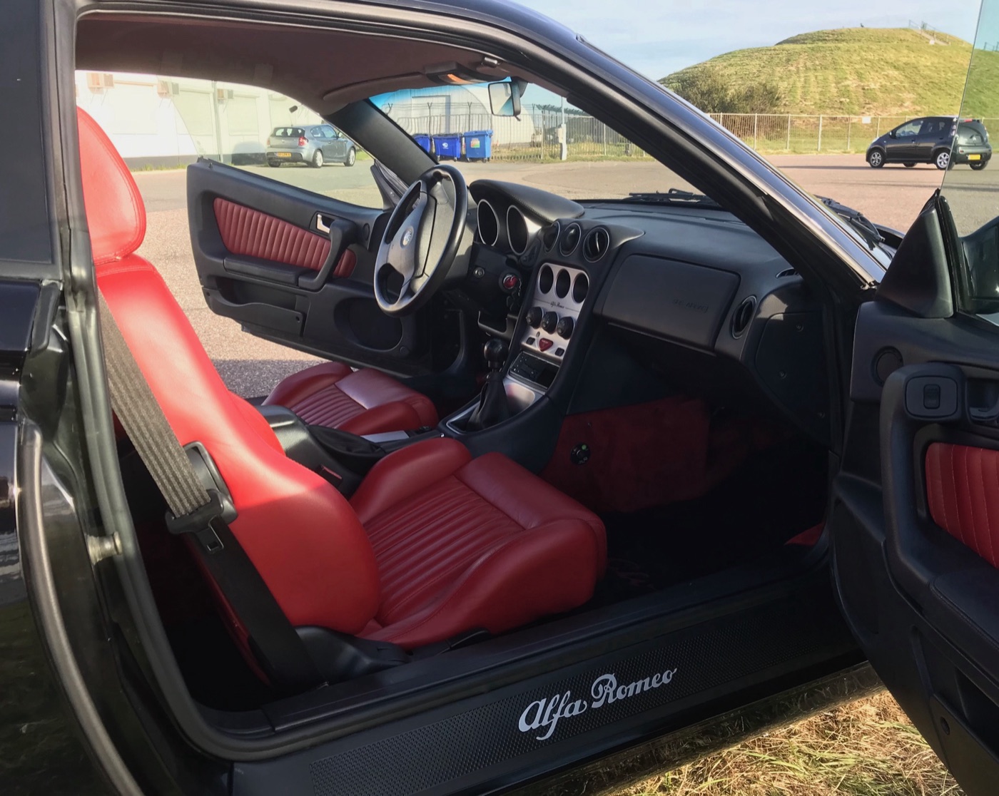 Interior of a black Alfa Romeo GTV seen through the open passenger door, with red pleated leather seats, red door inserts and a silver centre console