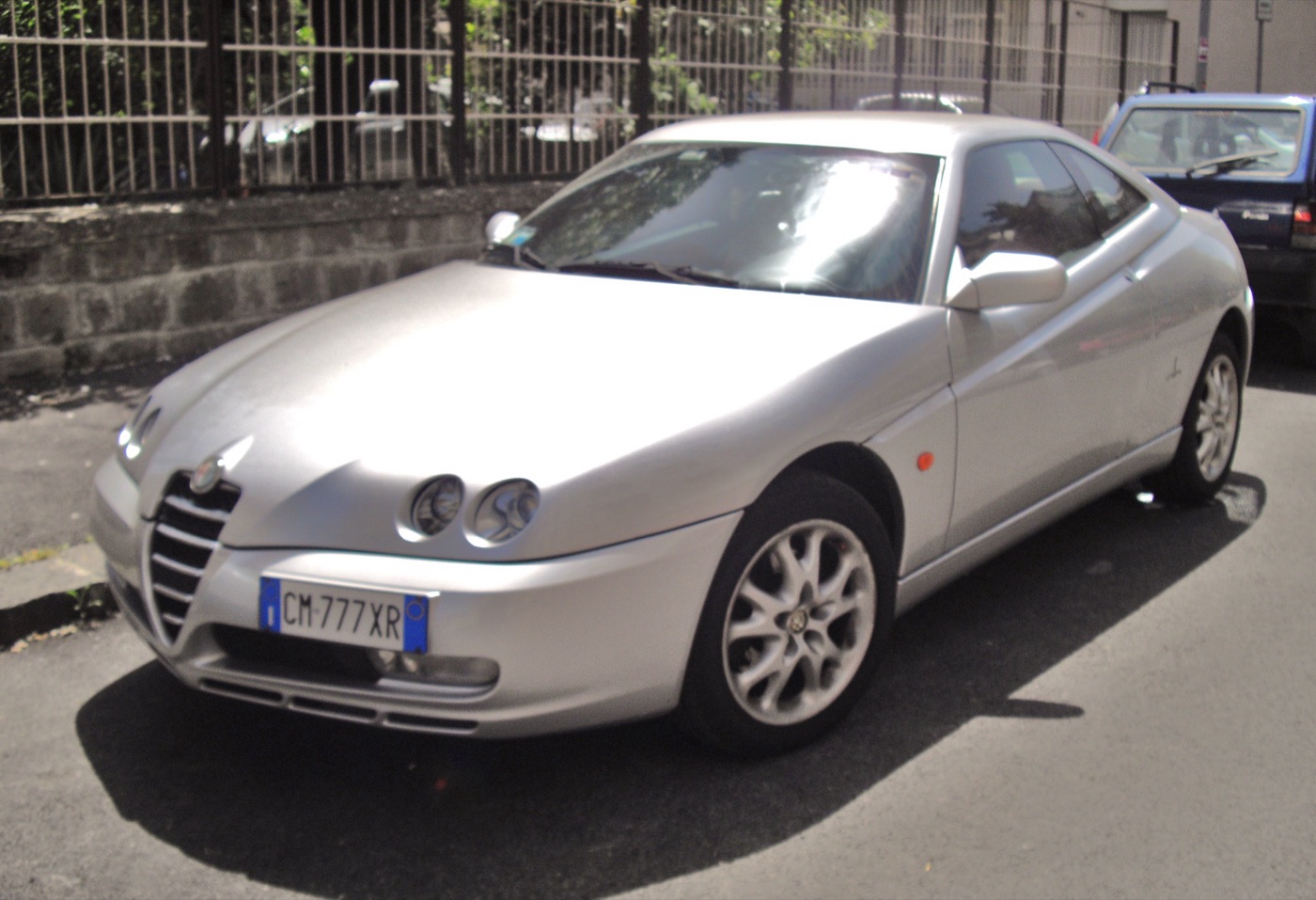 Silver Phase 3 Alfa Romeo GTV with a large chrome-barred grille and the number plate offset to the left of the bumper, parked on a street