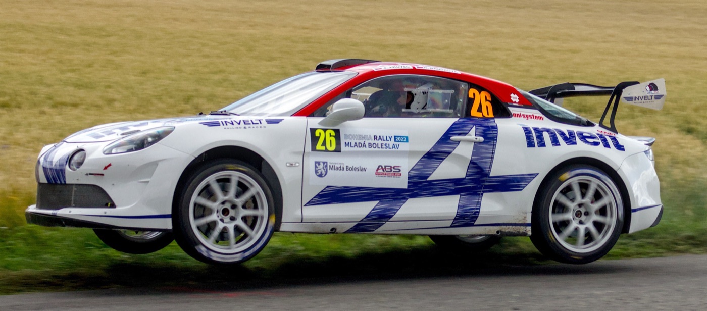 White and blue Alpine A110 Rally with a rear wing, wheels off the ground on a tarmac stage