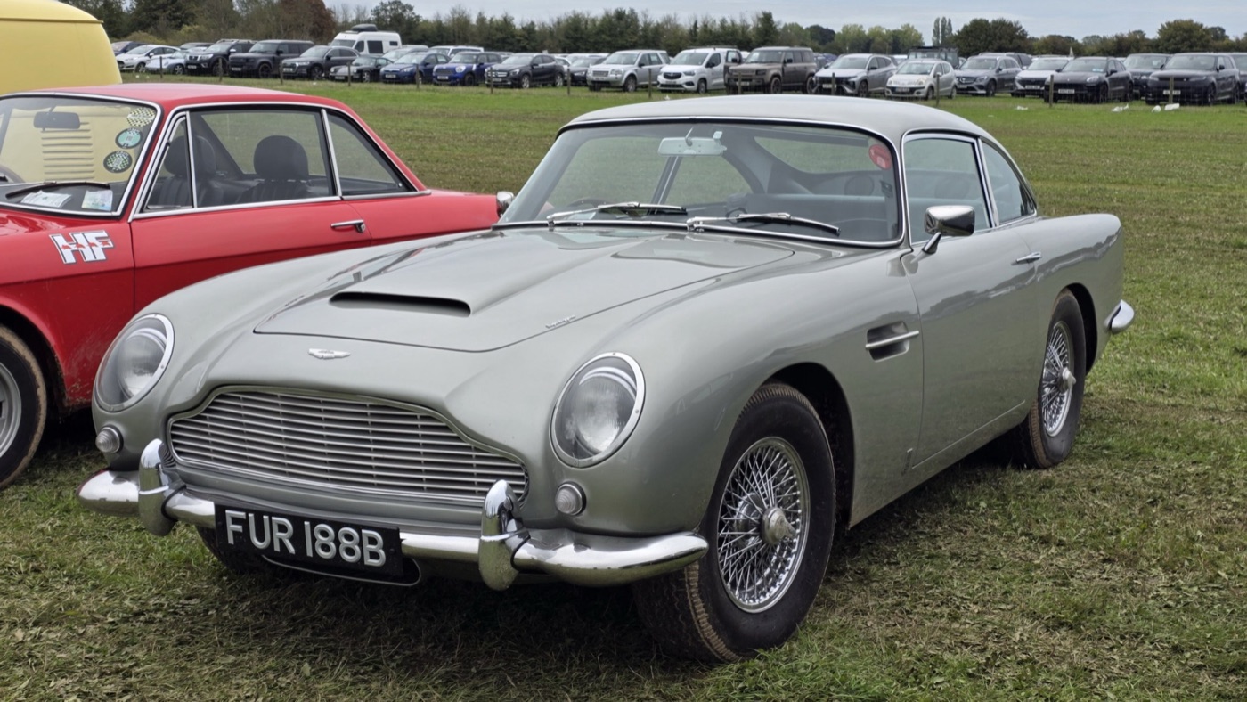 Silver 1964 Aston Martin DB5 saloon on grass at a car show, showing the covered headlamps, bonnet scoop and wire wheels