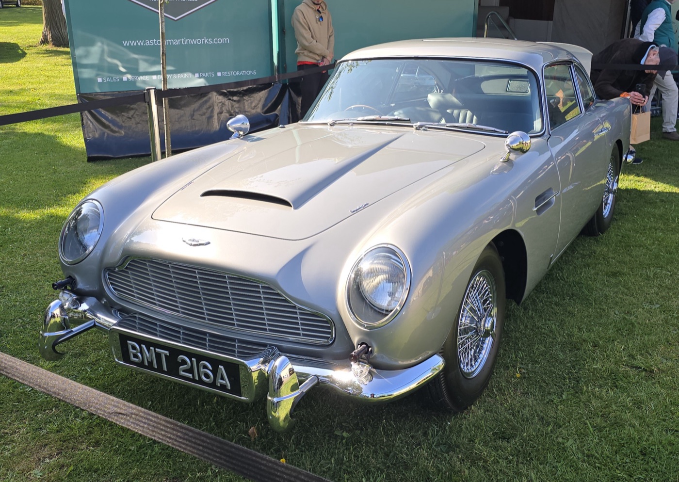 Silver Goldfinger Continuation DB5 with BMT 216A plates and over-rider rams, on grass in front of an Aston Martin Works stand