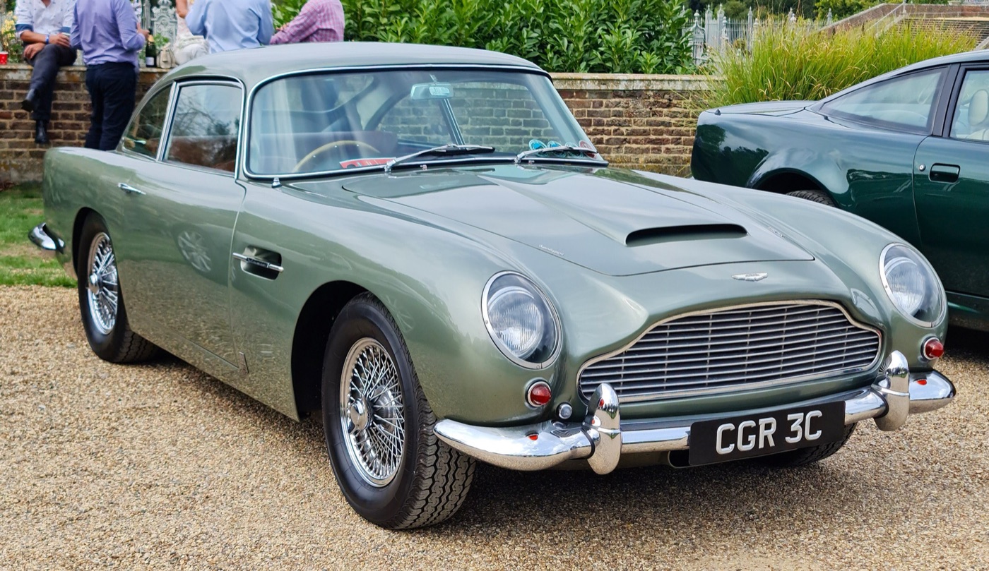 Sage-green DB5 saloon on gravel at a concours, front three-quarter view with wire wheels and chrome over-riders