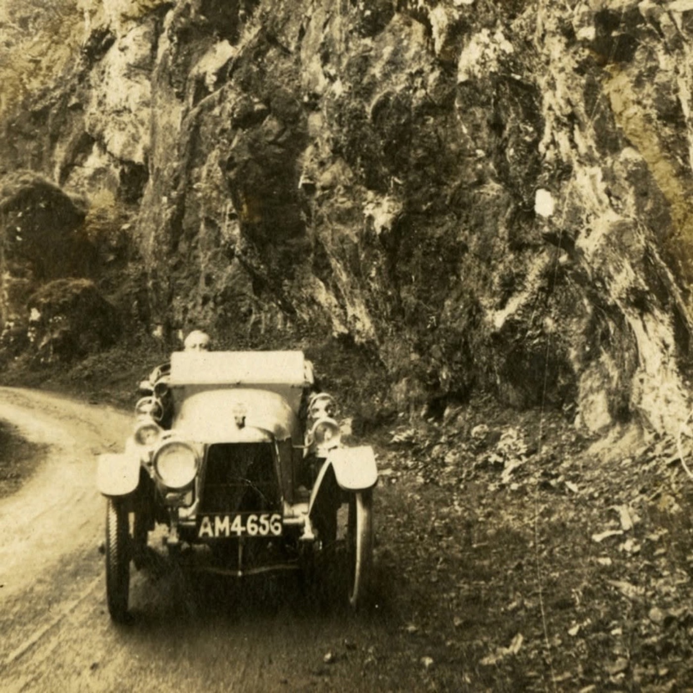 Sepia photograph of the 1915 Aston Martin prototype, registration AM 4656, on a narrow road below a rock face