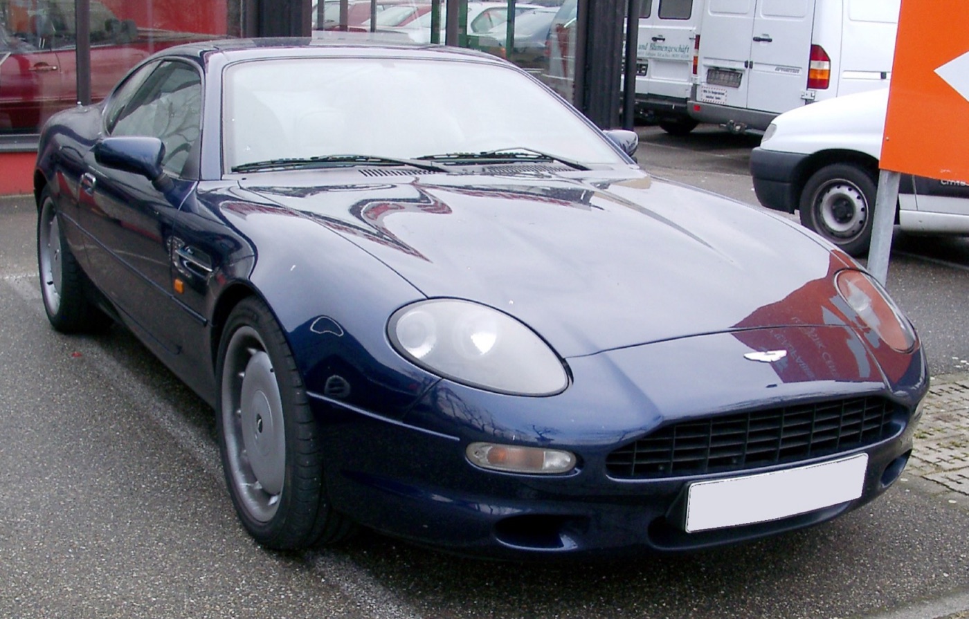 Dark blue Aston Martin DB7 coupé parked in front of a showroom with vans behind