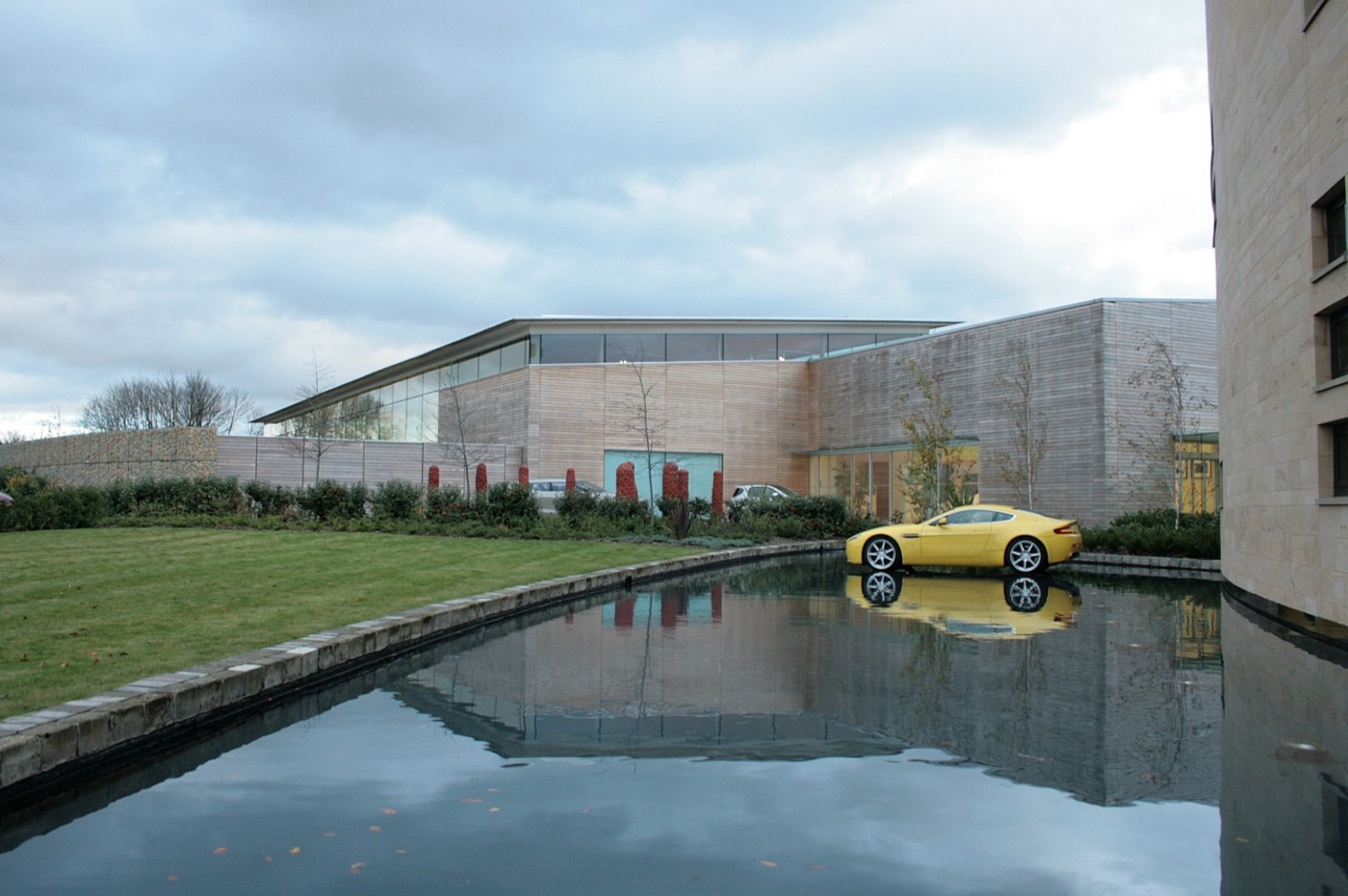Yellow Aston Martin V8 Vantage on a shallow pool in front of the timber-clad Aston Martin headquarters at Gaydon