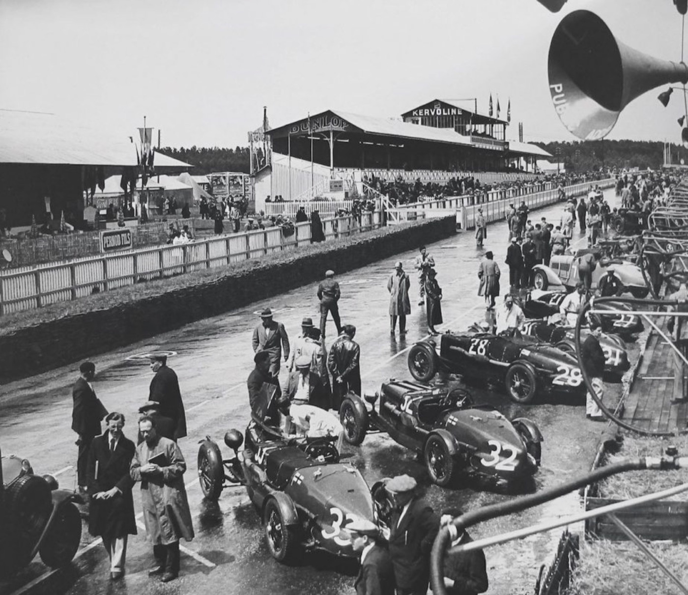Black and white photograph of Aston Martin Ulster racing cars numbered 28, 32 and 33 in the pits at Le Mans, with the Dunlop grandstand behind