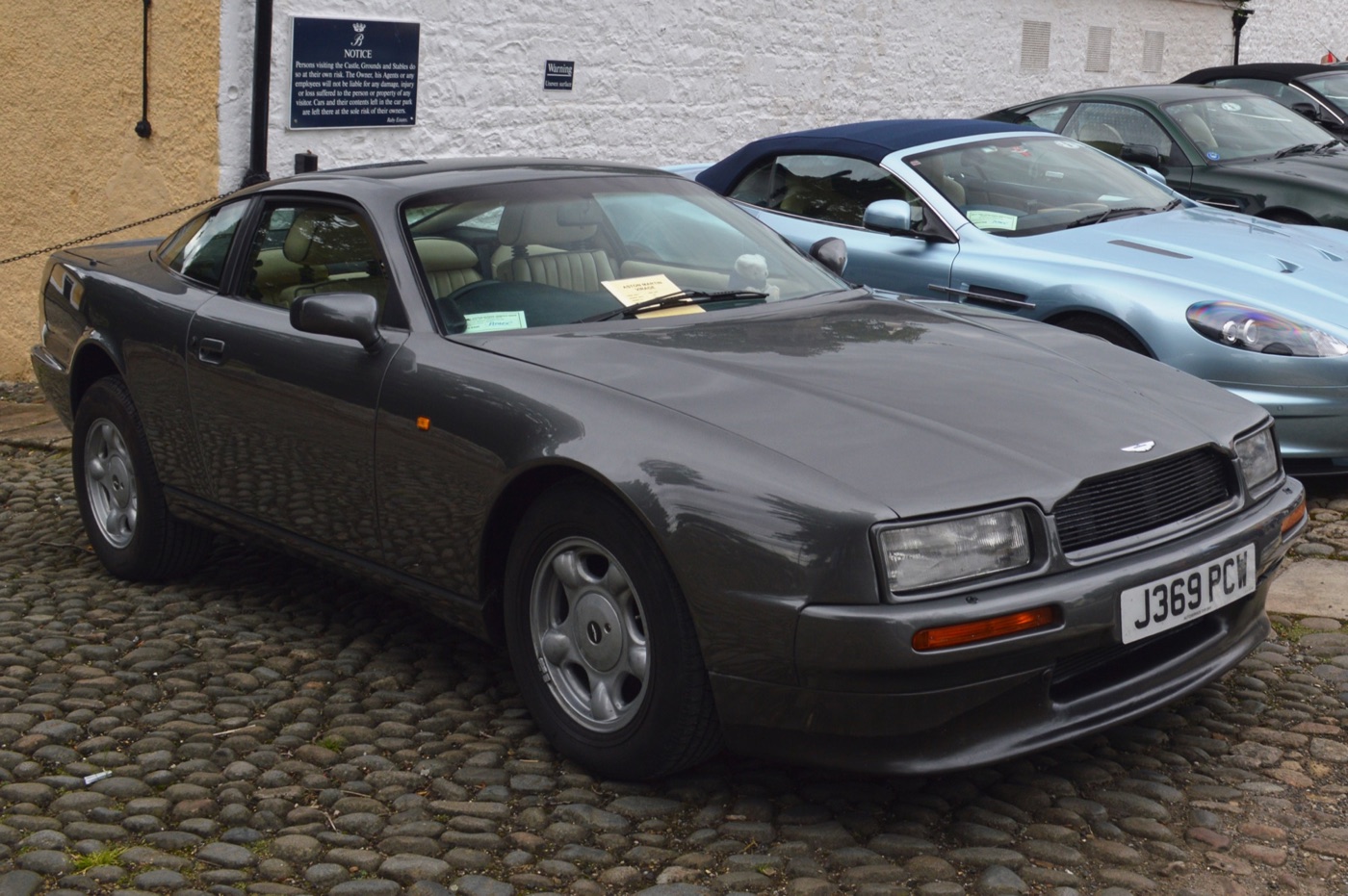 Grey 1991 Aston Martin Virage coupe on cobbles, with its slim Audi-sourced headlamps and body-coloured bumpers, beside a later Aston Martin convertible