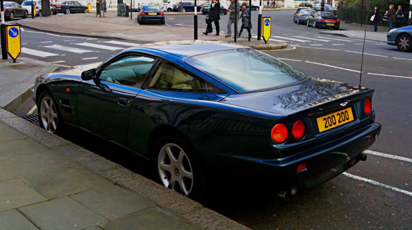Dark blue Aston Martin V8 Coupe seen from the rear quarter with four round tail lamps, parked on a London street