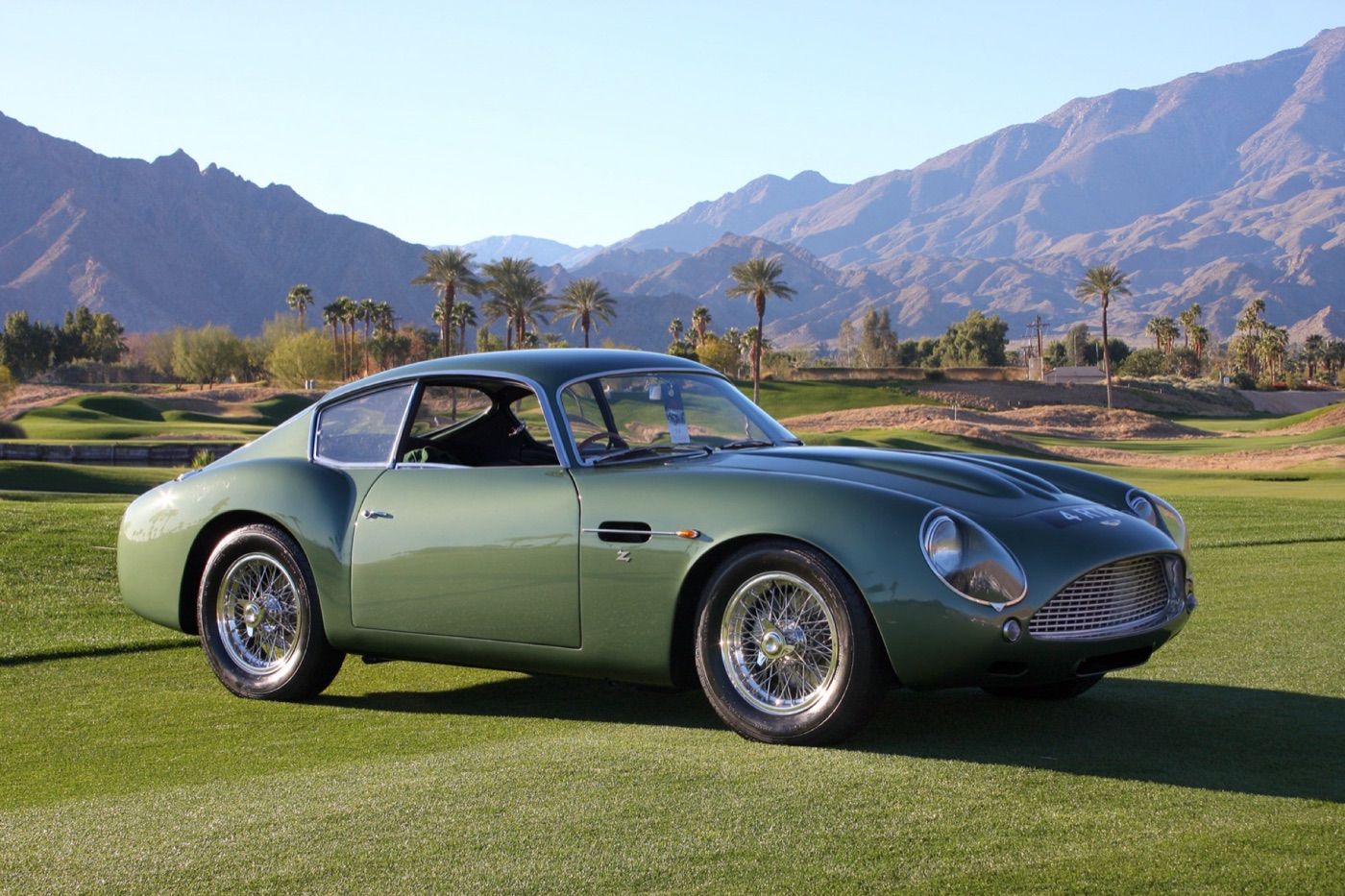 Green 1961 Aston Martin DB4 GT Zagato with wire wheels on a lawn, palm trees and desert mountains behind