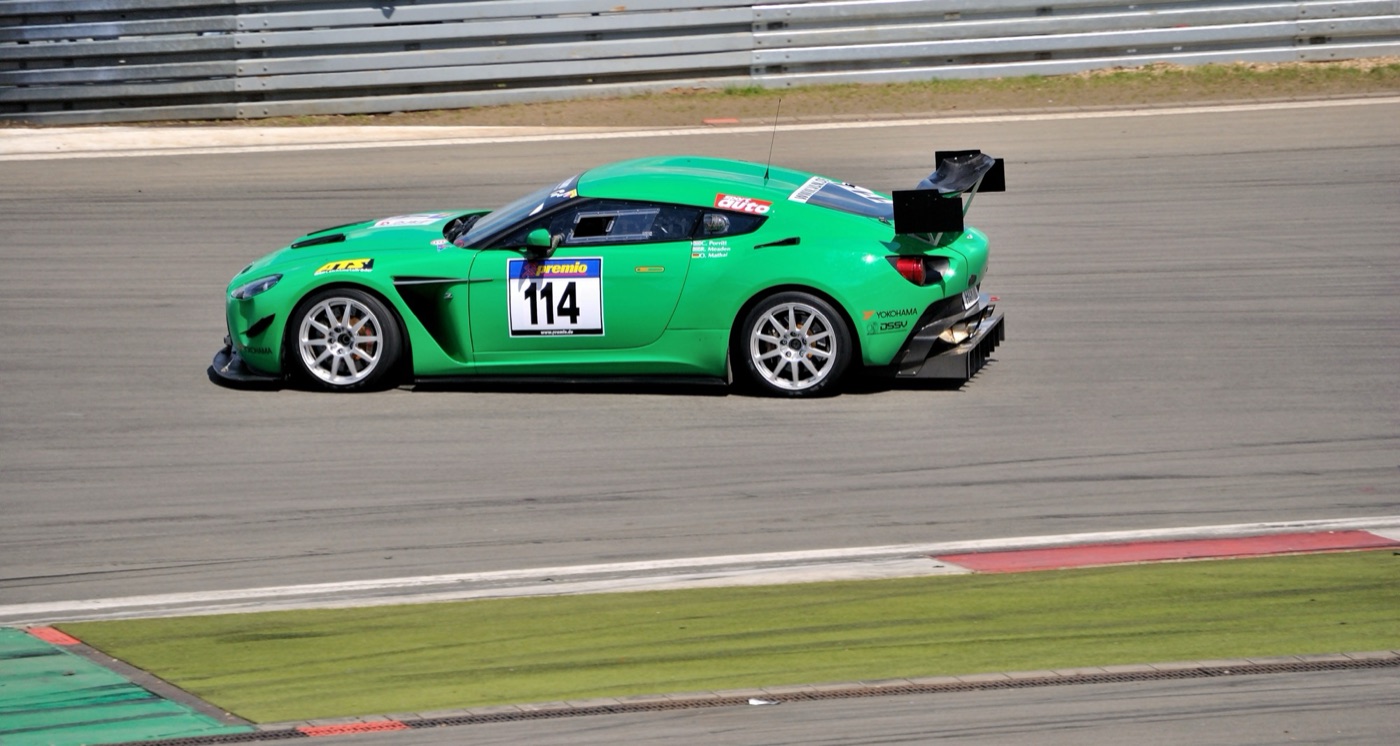 Green Aston Martin V12 Zagato race car with number 114, a rear wing and white wheels, at speed on the Nürburgring grand prix circuit
