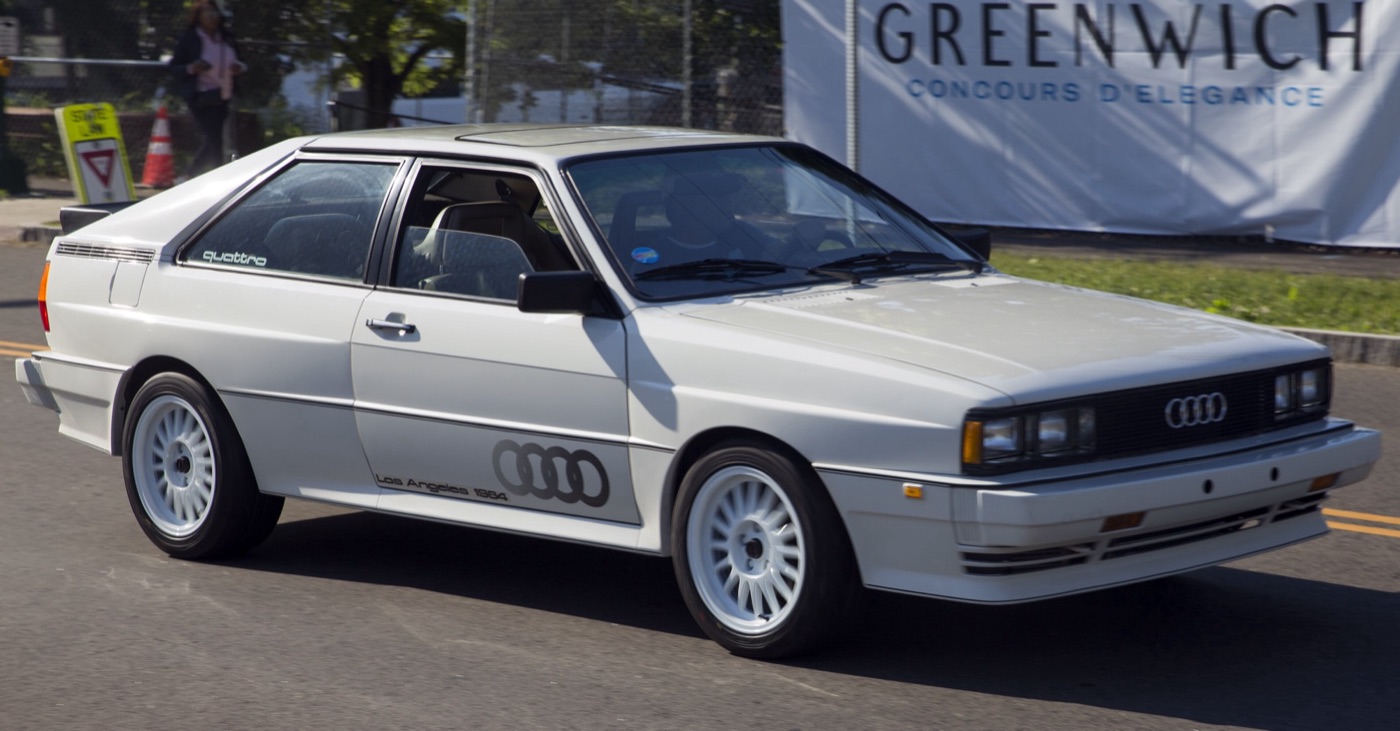 White 1984 Audi Quattro with Los Angeles 1984 graphics and four-ring logos on the doors, driving past a Greenwich Concours banner