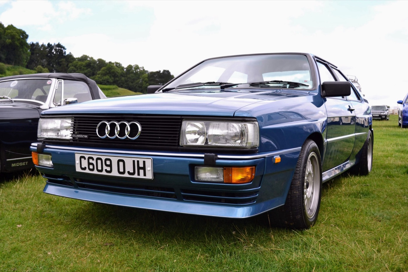 Front of a blue 1986 Audi Quattro with a black grille, four-ring badge and British number plate, parked on grass
