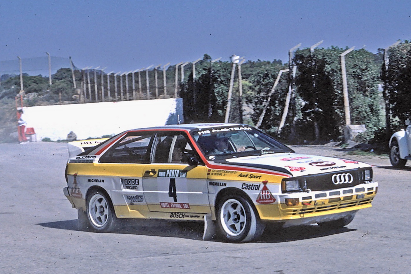 Yellow and white HB Audi Team Quattro A2 rally car number 4 sliding on a tarmac road near a fence on the 1984 Rally of Portugal