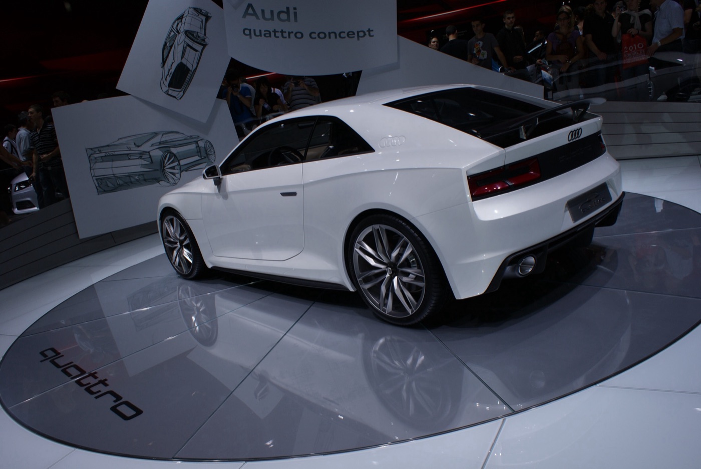 White Audi quattro concept coupé seen from the rear three-quarter on a turntable at the 2010 Paris Motor Show, with design sketches behind
