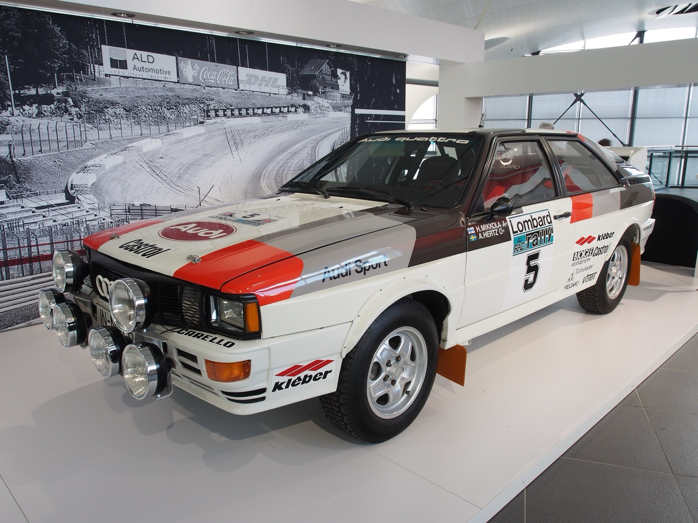 White Audi Quattro rally car with red and grey Audi Sport stripes, Lombard RAC Rally number 5 and a bank of extra lamps, on display indoors