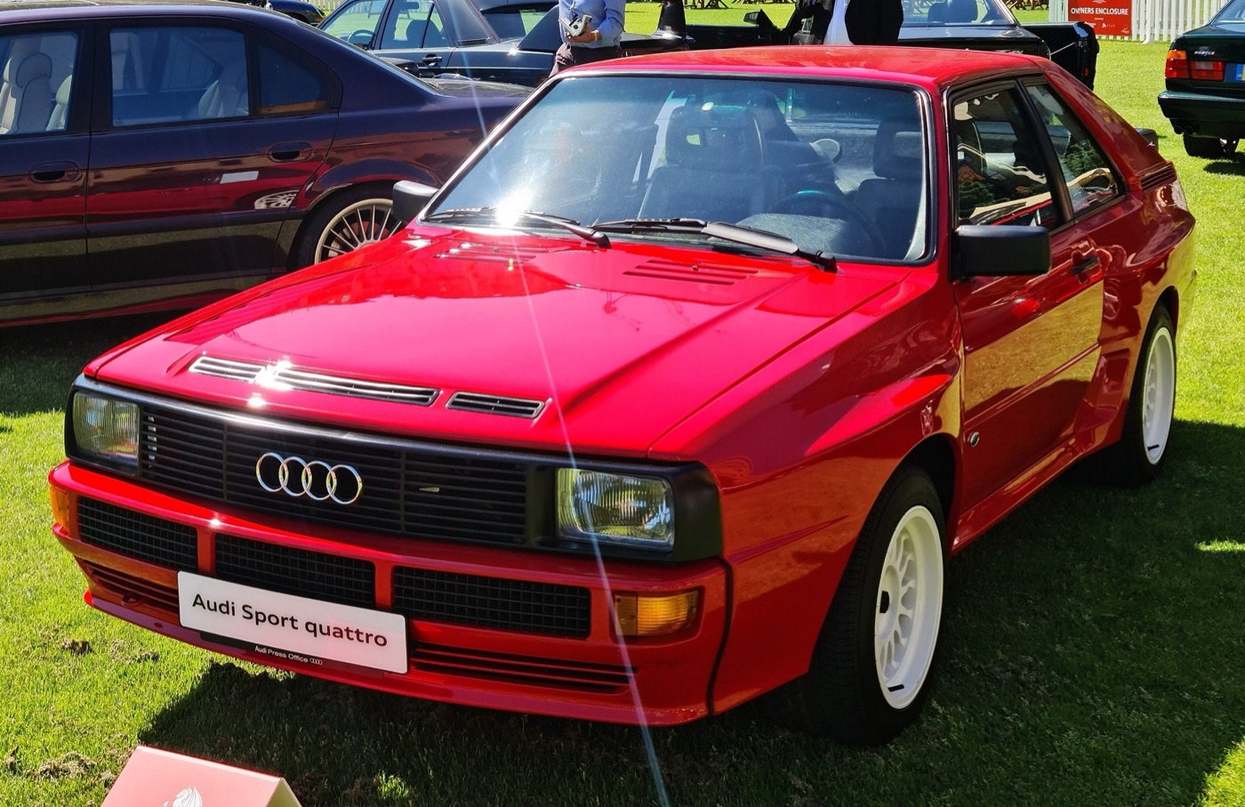 Red Audi Sport quattro with white wheels and bonnet vents parked on grass at a concours