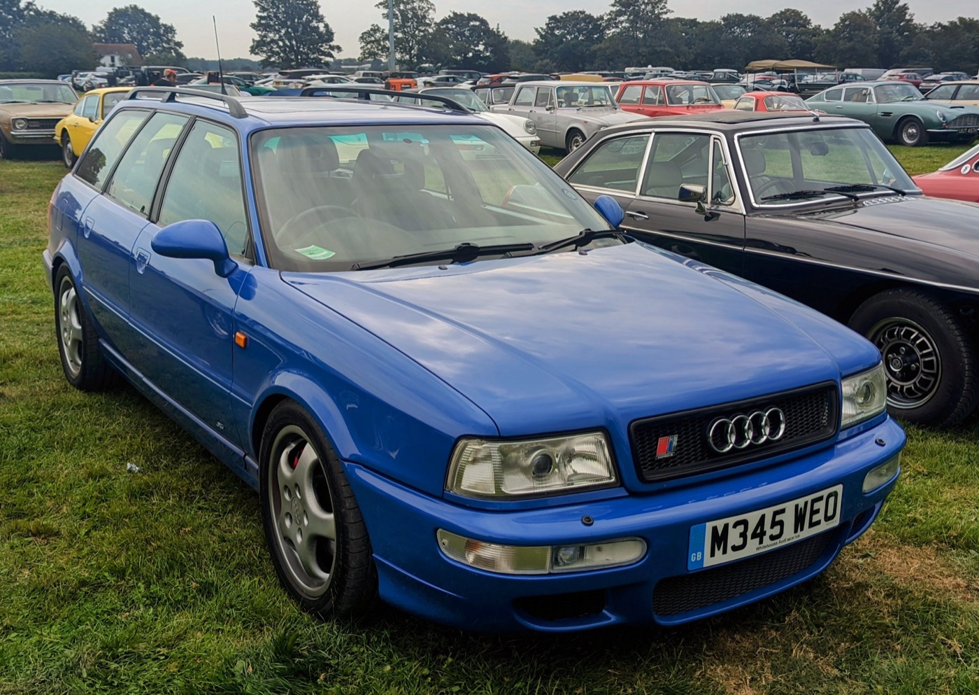 Blue Audi RS2 with roof rails, Porsche Cup wheels and a British number plate, parked on grass among classic cars