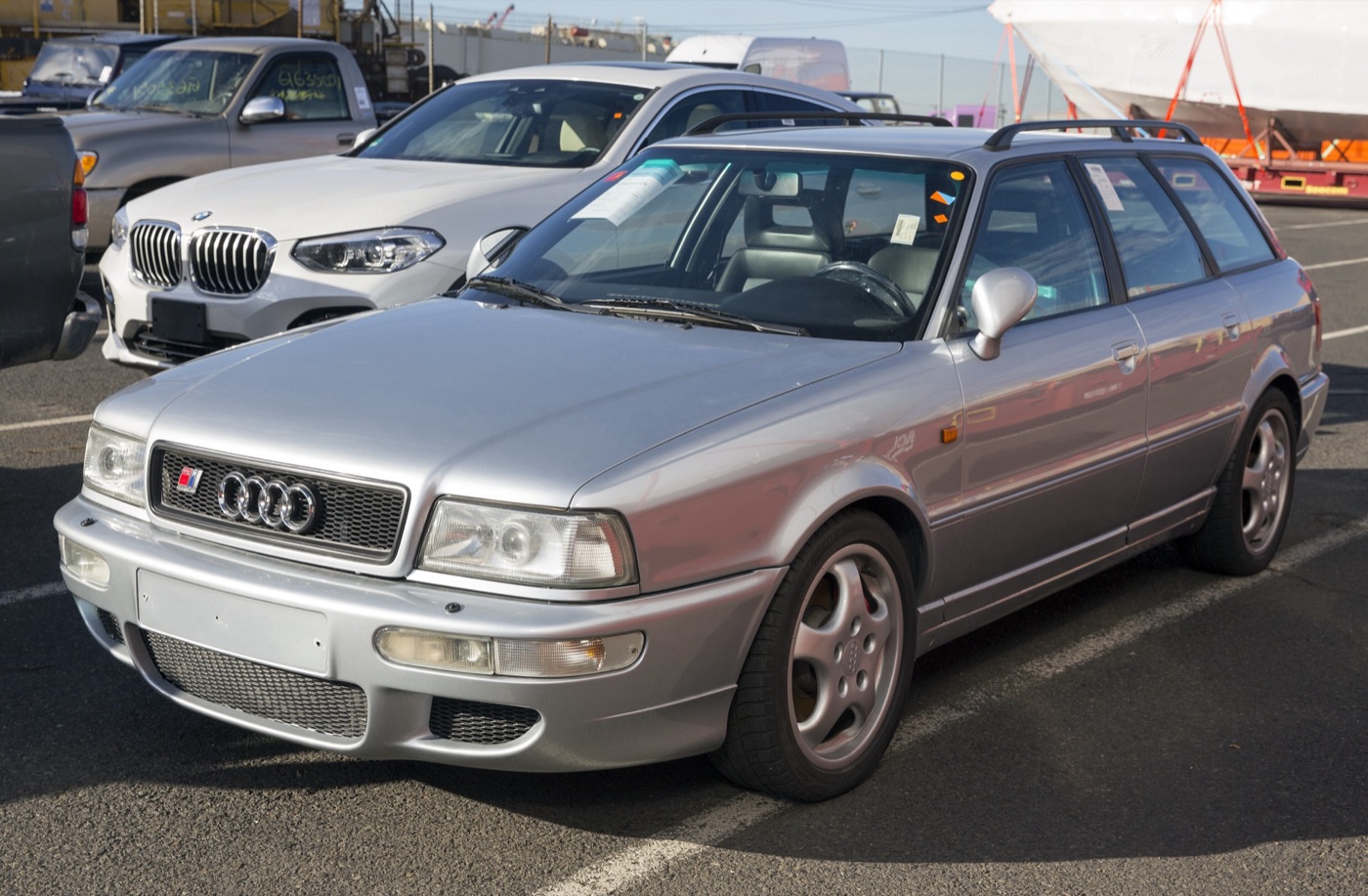 Silver Audi RS2 with stickers on its windscreen in a port vehicle compound, beside a white BMW
