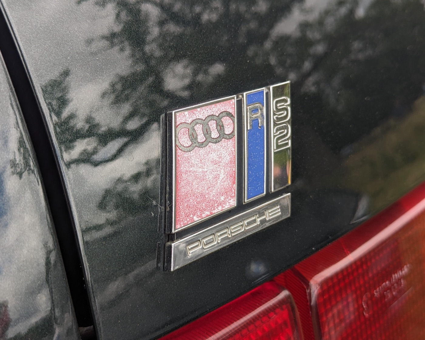 Close-up of the Audi RS2 badge on a dark tailgate: the four rings, a blue R, a chrome S2 and a bar reading Porsche