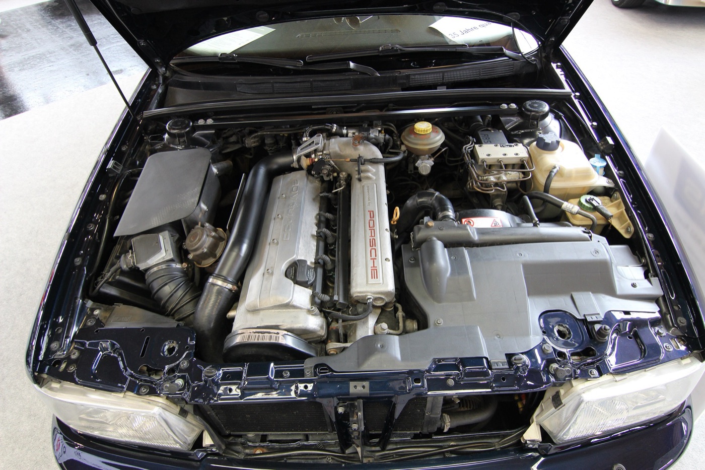 Engine bay of an Audi RS2 with its silver five-cylinder cam cover marked Porsche, the intercooler pipe and the bonnet open