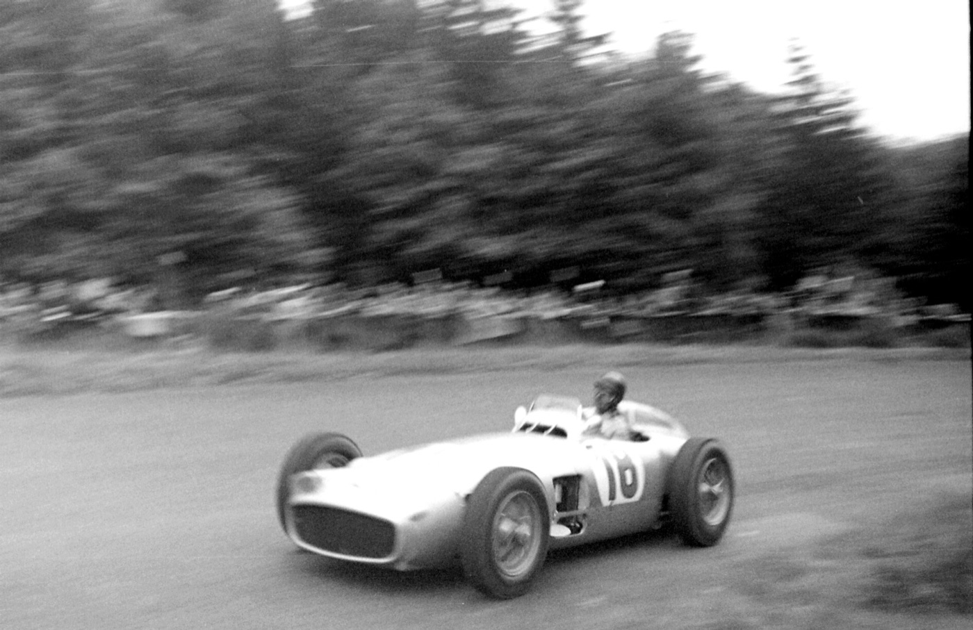 Black and white panning photograph of Juan Manuel Fangio in the silver Mercedes-Benz W196, number 18, at speed on the Nürburgring with trees behind