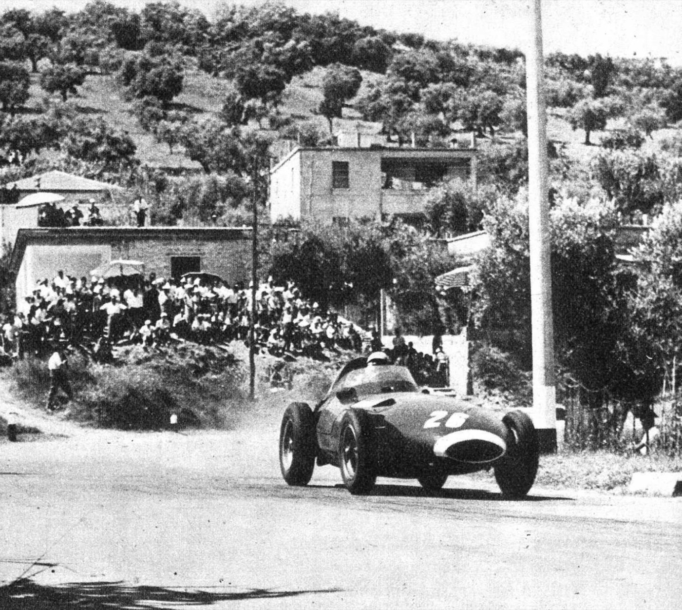 Black and white photograph of Stirling Moss’s Vanwall, number 26, rounding a chicane on the Pescara street circuit with spectators on the bank behind