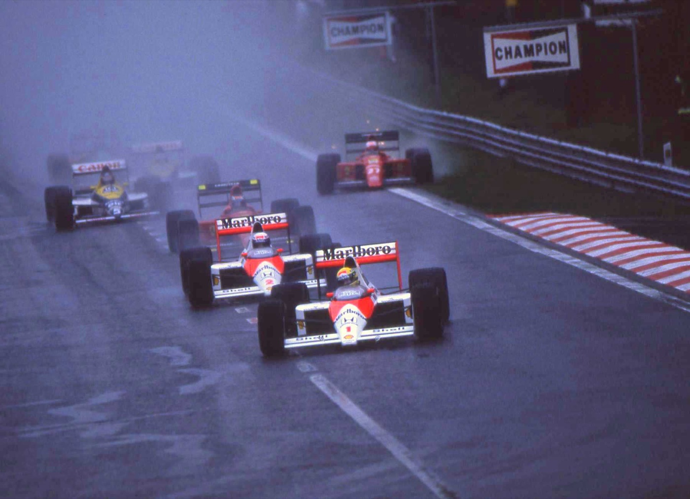 Two red and white McLaren-Honda cars lead the field through spray on a wet track at Spa, Senna in front with his yellow helmet and Prost behind