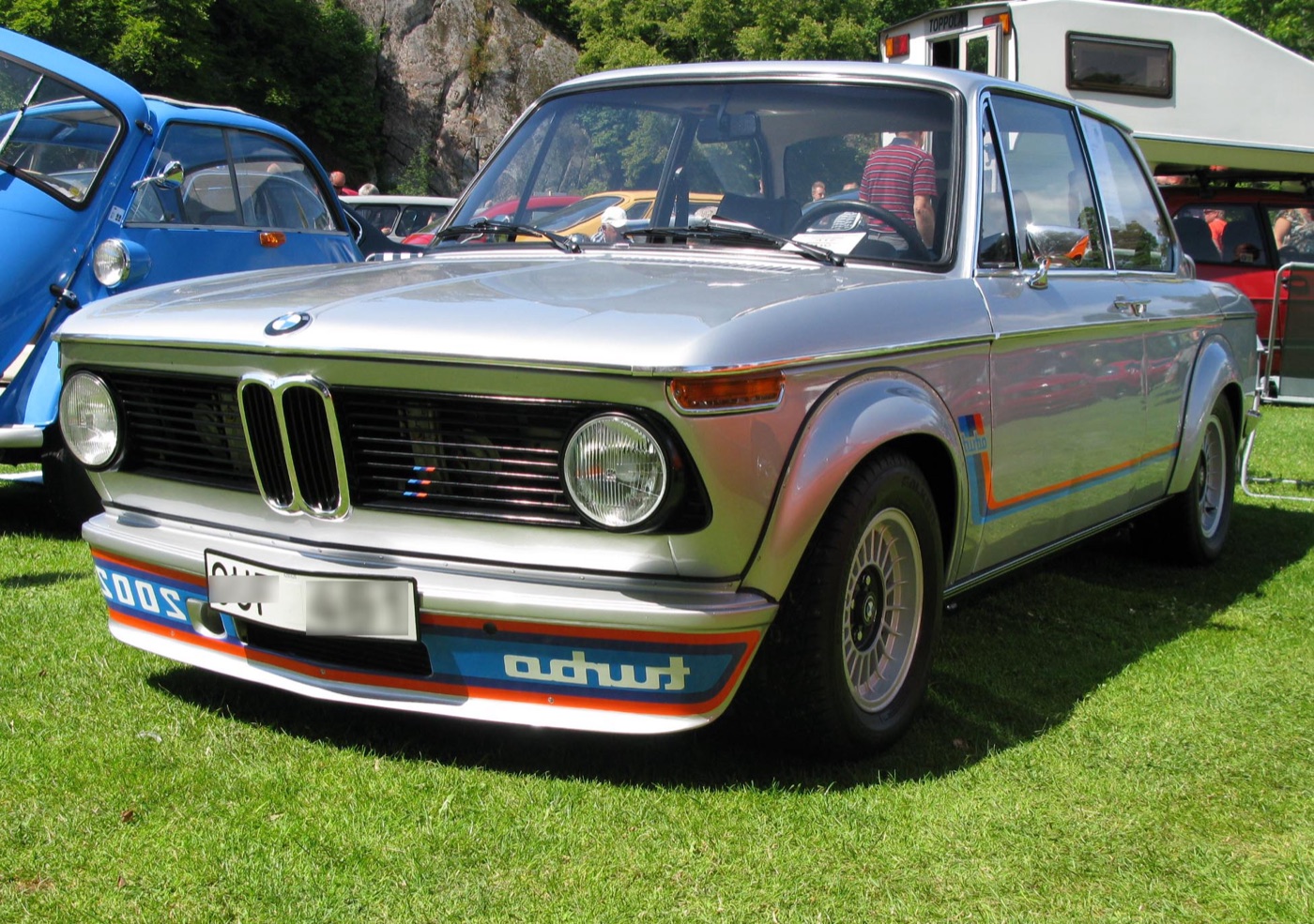 Silver 1974 BMW 2002 turbo with reversed turbo lettering on its front spoiler, parked on grass at a car show