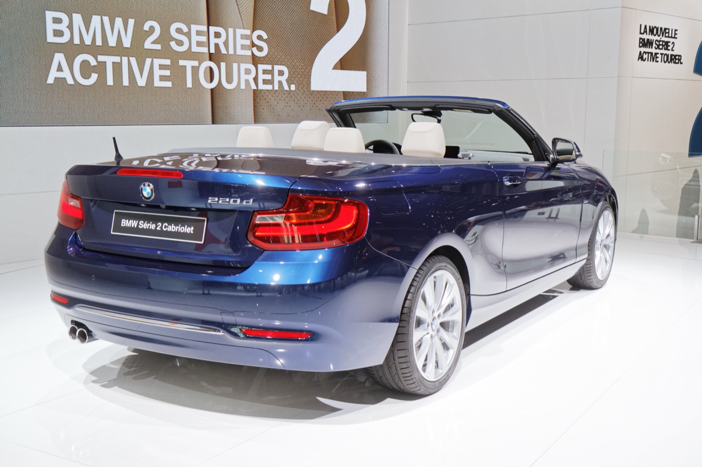 Dark blue BMW 220d Convertible with its fabric roof folded, seen from the rear on a motor show stand