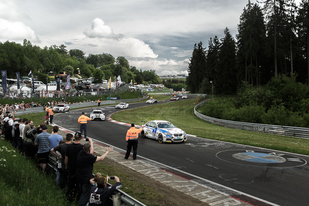 BMW M235i Racing number 308 leading cars through a corner on the Nürburgring Nordschleife with spectators behind the fence