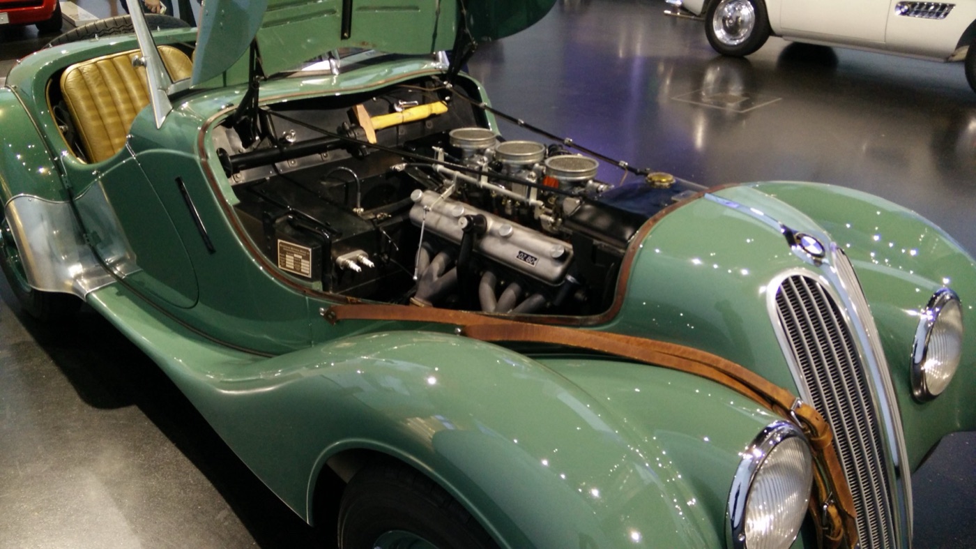 Engine bay of a green BMW 328 roadster with the bonnet side raised, showing three downdraught carburettors and the six-branch exhaust manifold