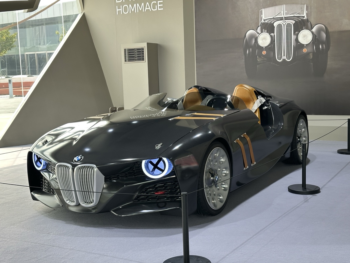 Dark grey BMW 328 Hommage roadster with tan leather straps and seats, behind a rope, a photograph of a 1930s 328 on the wall