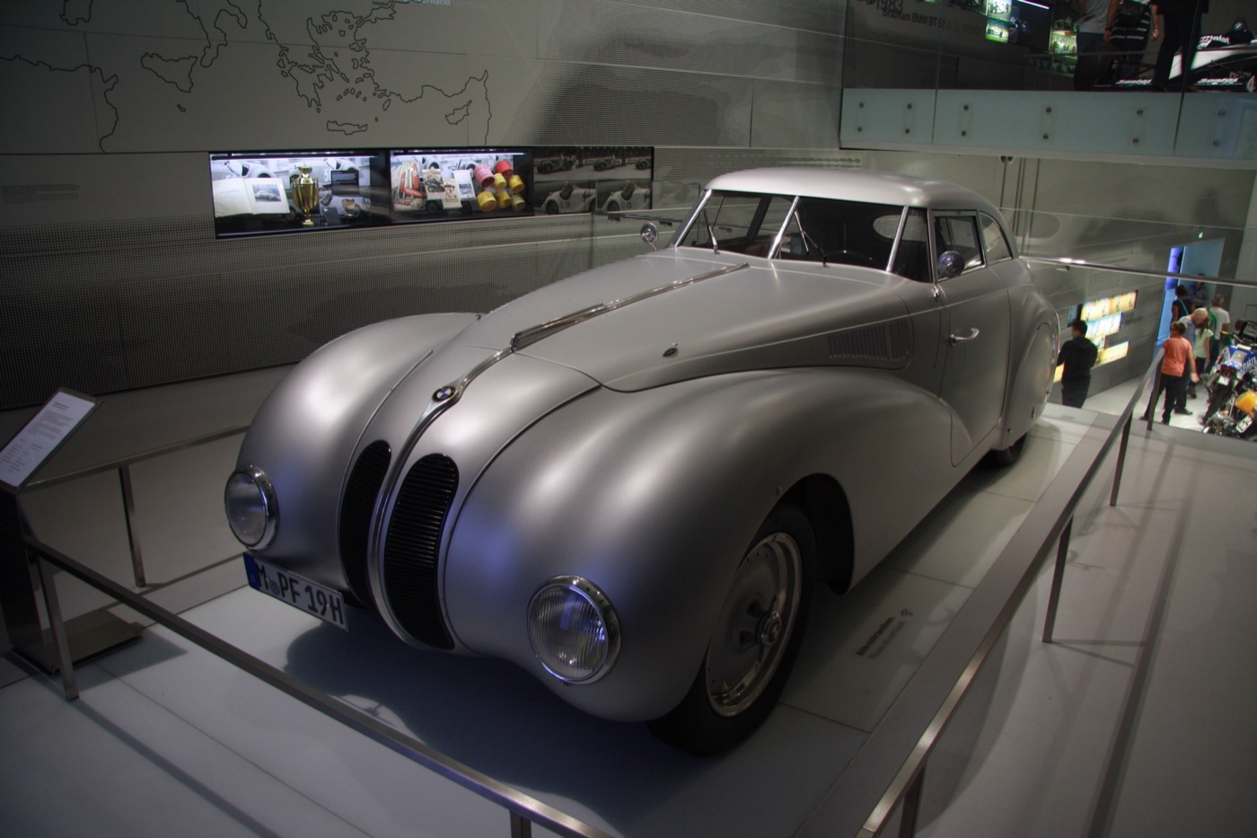 Silver fastback BMW 328 Kamm coupé reconstruction with a long bonnet on a museum plinth behind a steel rail