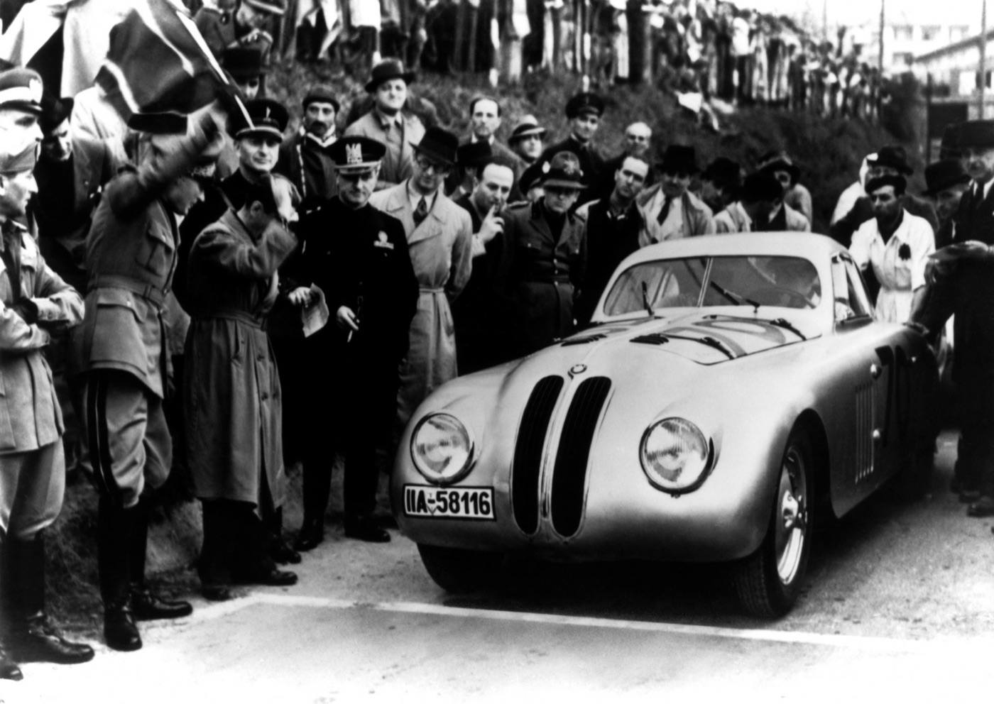 Black and white photograph of the silver BMW 328 Touring coupé with German plate IIA-58116, surrounded by officials, uniformed men and spectators