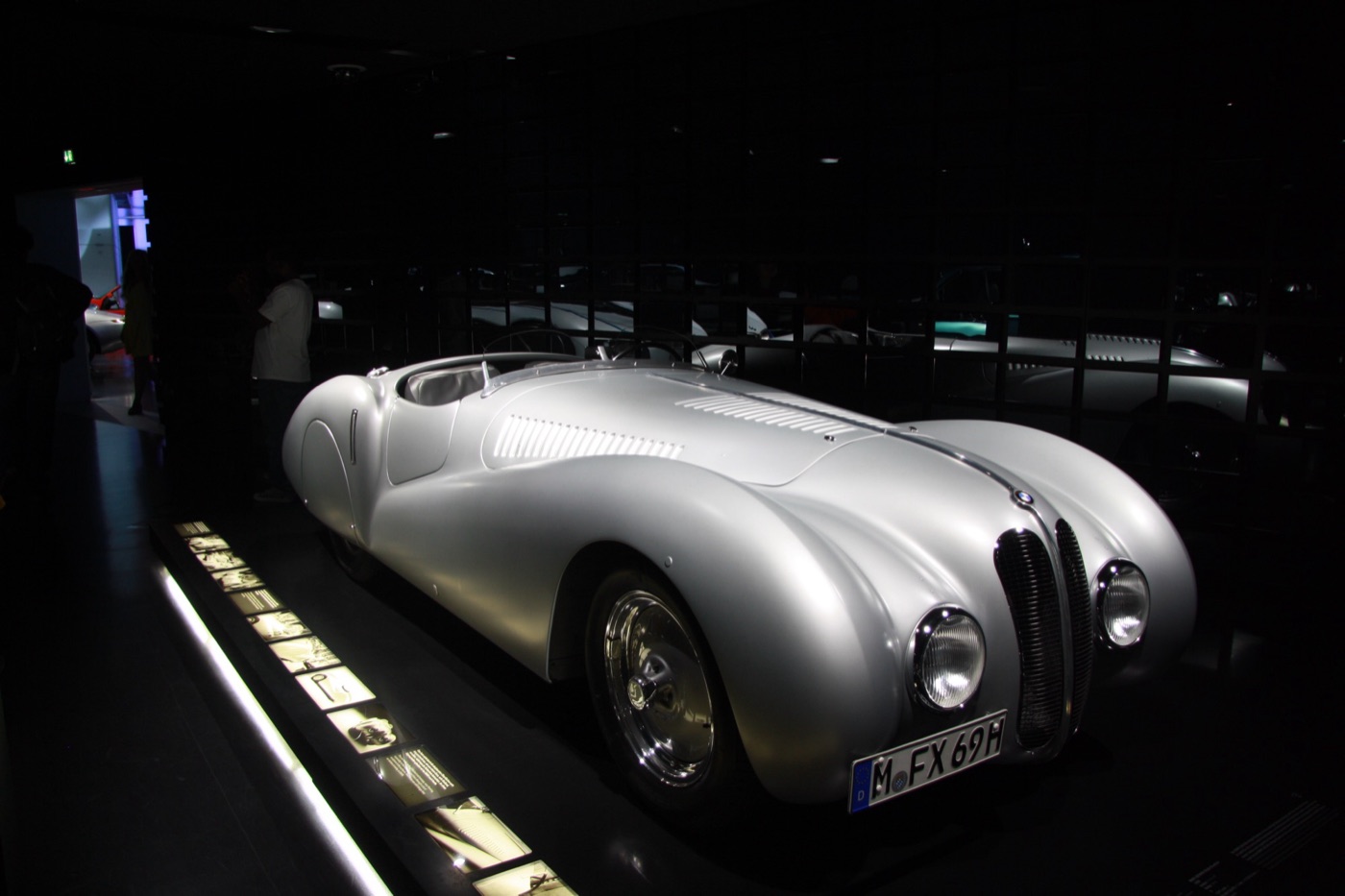 Silver streamlined BMW 328 Mille Miglia roadster with louvred bonnet and twin-kidney grille under a spotlight in a dark museum hall