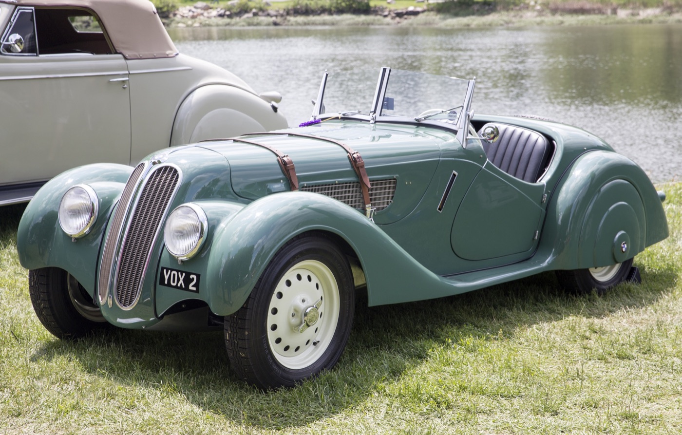 Green 1937 BMW 328 roadster on grass beside a river, with a narrow twin-kidney grille, headlamps faired into the wings, two leather bonnet straps and cream centre-lock wheels