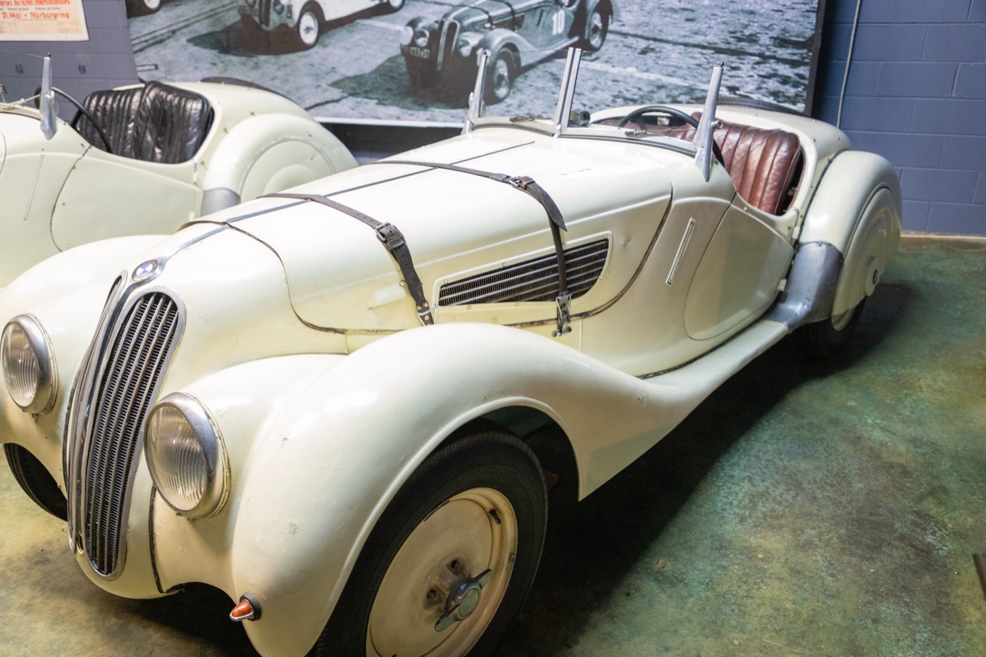 Ivory-white unrestored BMW 328 roadster with worn paint, leather bonnet straps and brown seats in a museum, a period race photograph behind