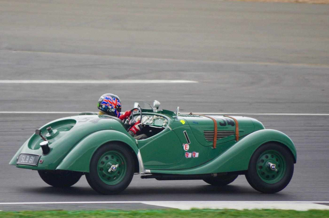 Green BMW 328 roadster with leather bonnet straps racing on the Silverstone circuit, its driver in a Union Jack helmet