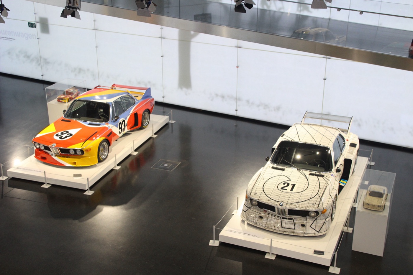 Two BMW 3.0 CSL Art Cars on white plinths in the BMW Museum: Calder’s in red, yellow and blue with number 93, and Stella’s black-and-white grid car with number 21, each with its scale model